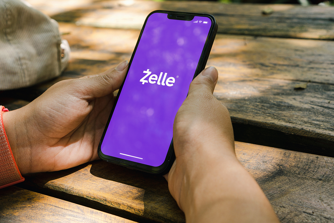 Person sitting at picnic table holding cellphone with Zelle pulled up on screen